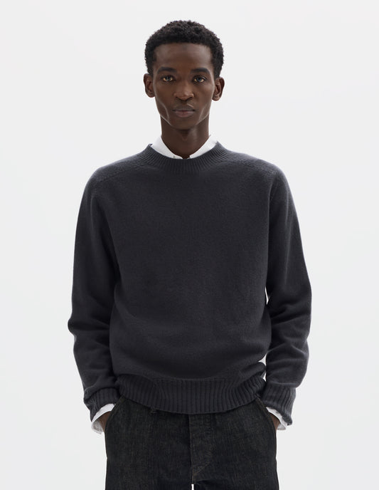 Cropped body image, front view of model wearing dark grey merino cashmere seamless jumper with ribbed crew neck, cuffs and hem. Styled over white shirt, and with black jeans.