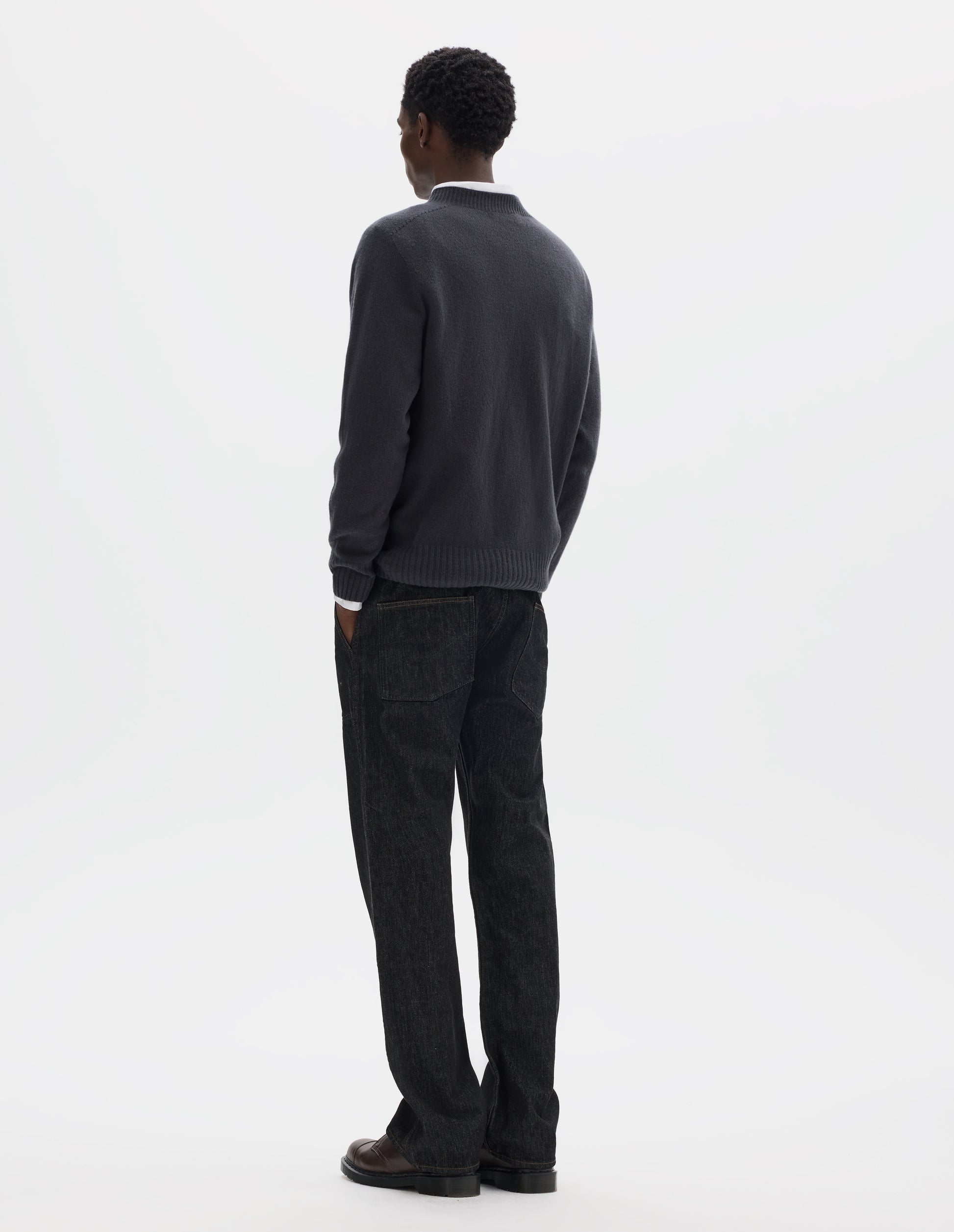 Full body image, back view of model wearing dark grey merino cashmere seamless jumper with ribbed crew neck, cuffs and hem. Styled over white shirt, and with black jeans and brown leather shoes.