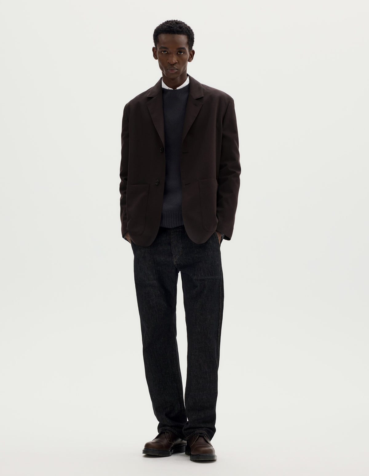 Full body image, front view of model wearing dark brown relaxed wool blazer with dropped shoulders, horn buttons and deep patch pockets. Styled over dark grey jumper and white shirt, and with black jeans and brown leather shoes.