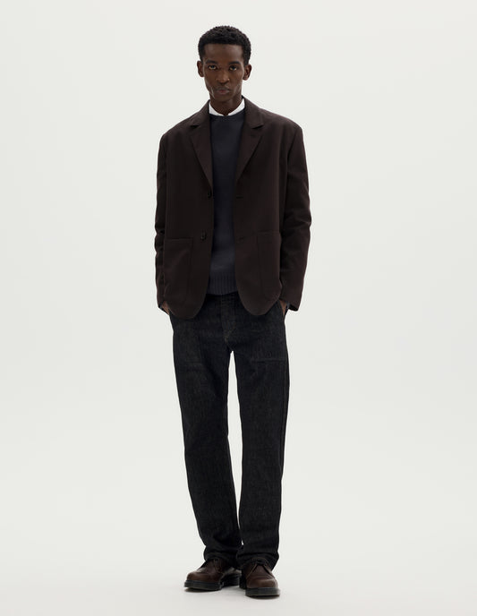Full body image, front view of model wearing dark brown relaxed wool blazer with dropped shoulders, horn buttons and deep patch pockets. Styled over dark grey jumper and white shirt, and with black jeans and brown leather shoes.