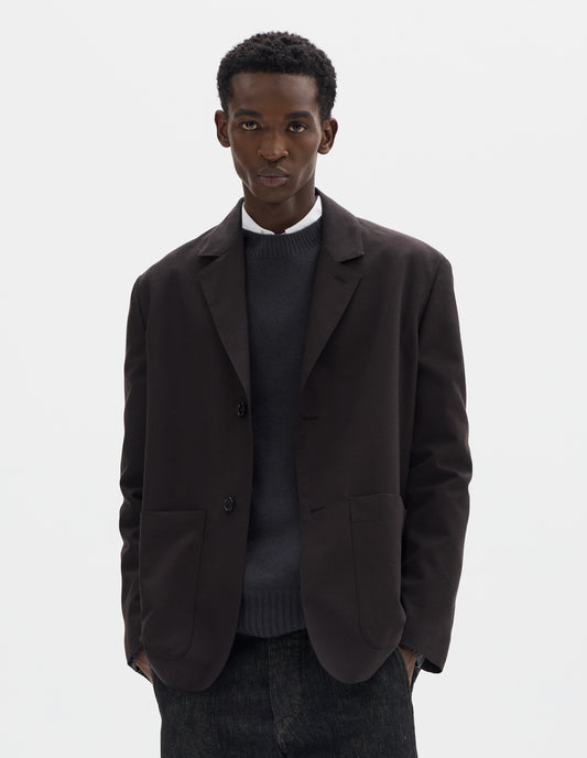 Cropped body image, front view of model wearing dark brown relaxed wool blazer with dropped shoulders, horn buttons and deep patch pockets. Styled over dark grey jumper and white shirt, and with black jeans.