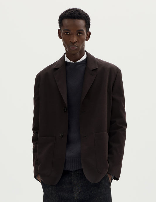 Cropped body image, front view of model wearing dark brown relaxed wool blazer with dropped shoulders, horn buttons and deep patch pockets. Styled over dark grey jumper and white shirt, and with black jeans.