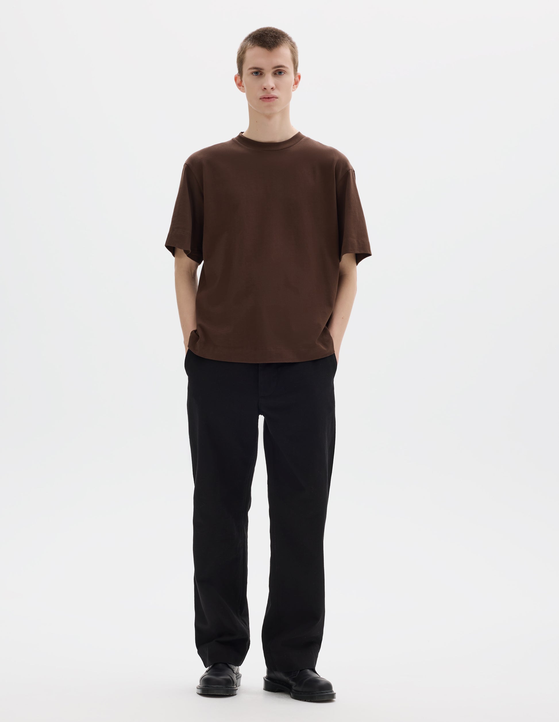 Full body image, front view of model wearing relaxed dark brown jersey t-shirt with crew neck and boxy cut. Styled with black trousers and black leather shoes.