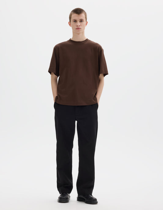 Full body image, front view of model wearing relaxed dark brown jersey t-shirt with crew neck and boxy cut. Styled with black trousers and black leather shoes.