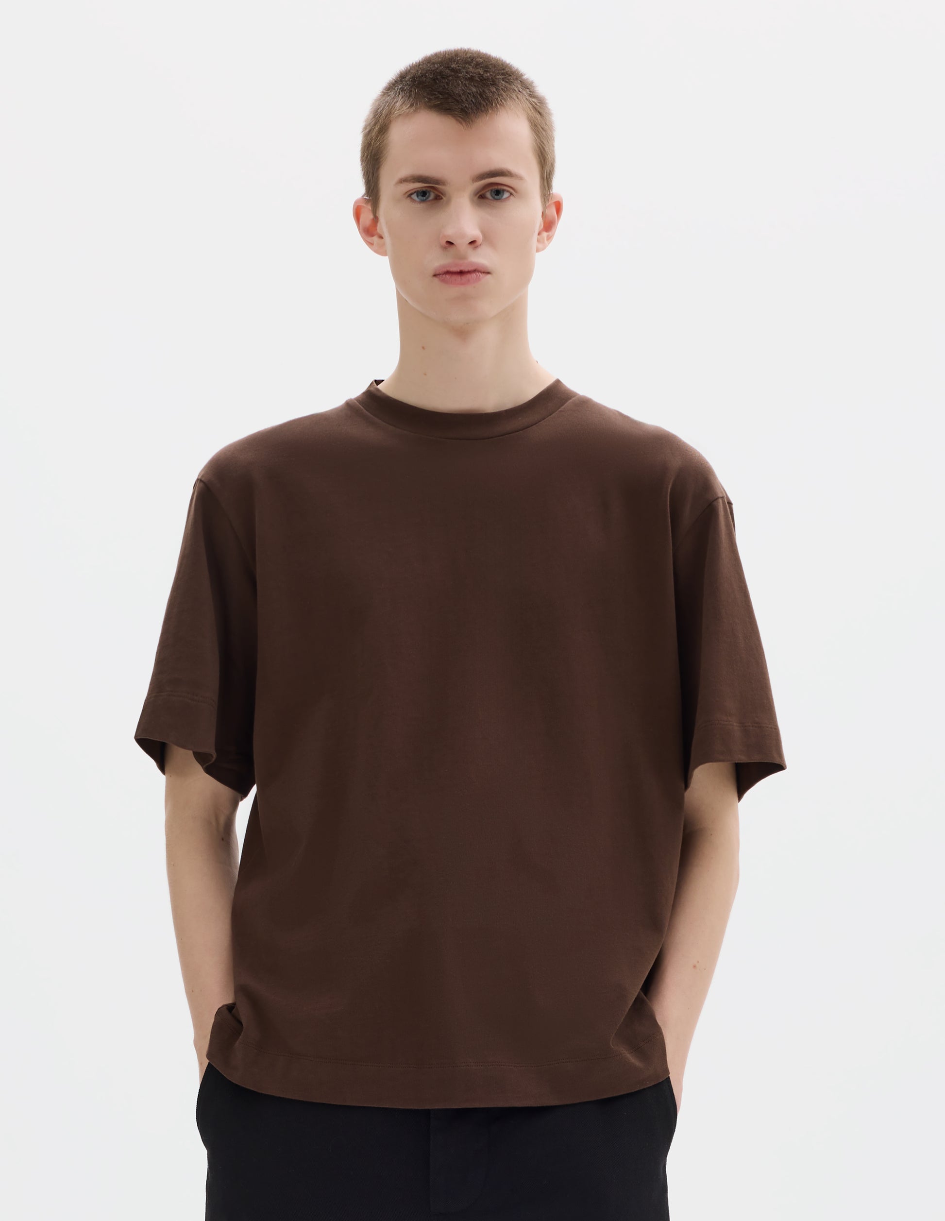 Cropped body image, front view of model wearing relaxed dark brown jersey t-shirt with crew neck and boxy cut. Styled with black trousers.