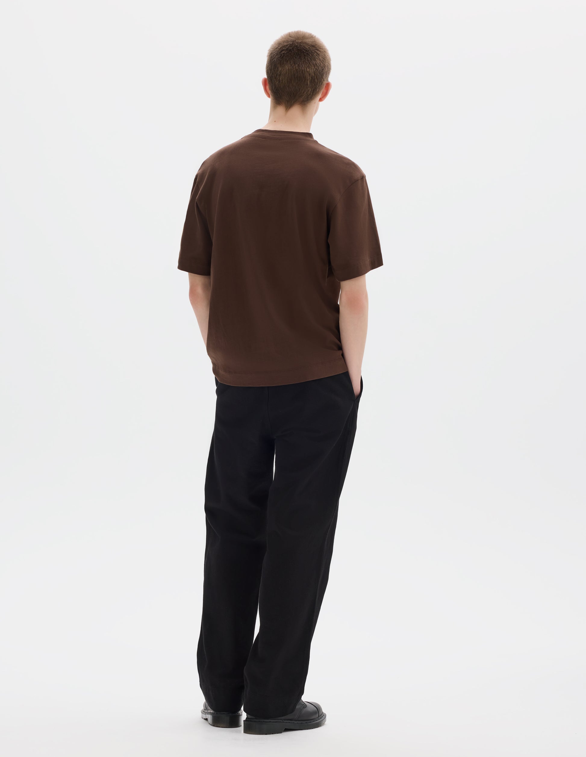 Full body image, back view of model wearing relaxed dark brown jersey t-shirt with crew neck and boxy cut. Styled with black trousers and black leather shoes.