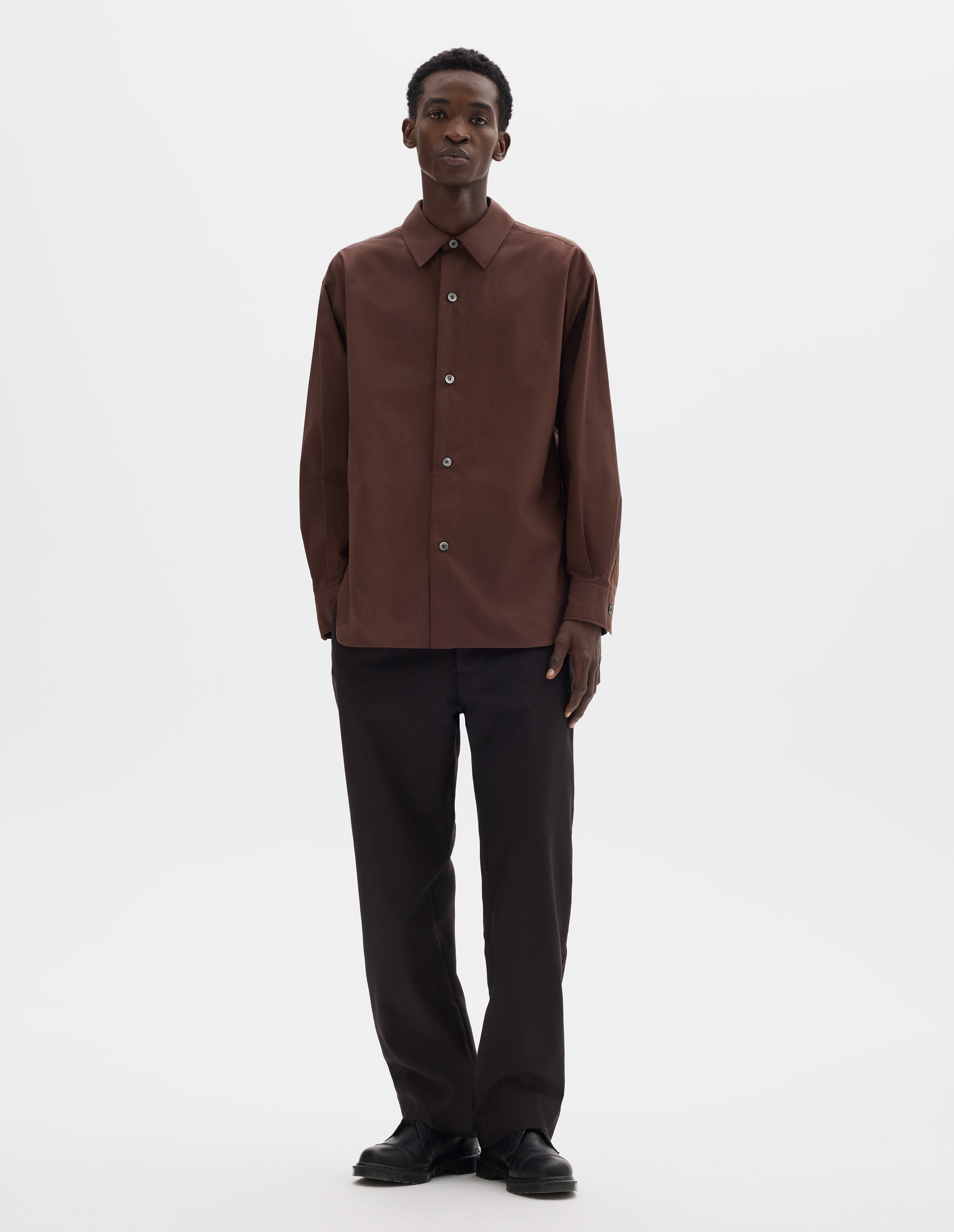 Full body image, front view of model wearing relaxed brown cotton shirt with mother of pearl buttons on wide stitch-through placket and curved hem. Styled with dark brown trousers and black leather shoes.
