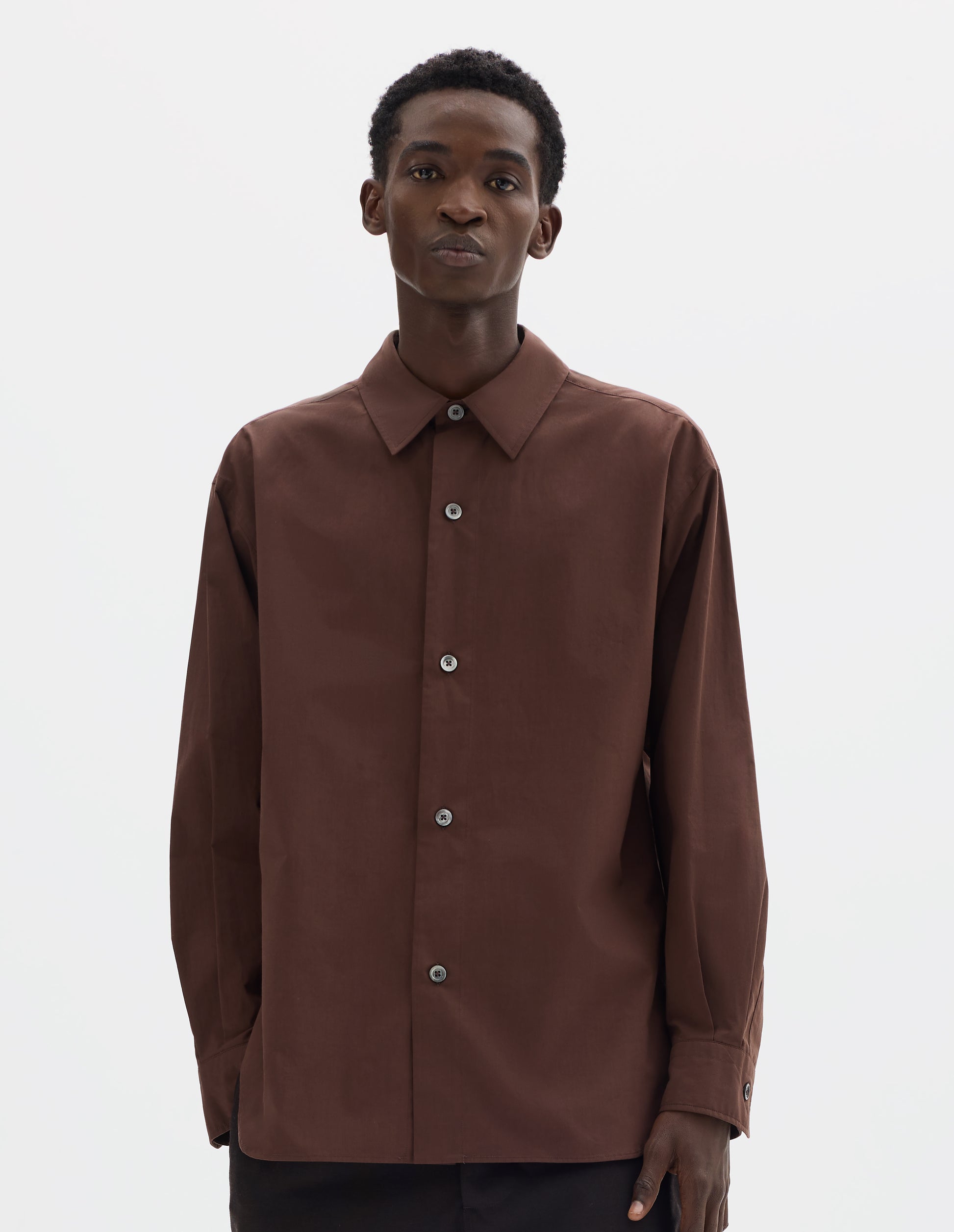 Cropped body image, front view of model wearing relaxed brown cotton shirt with mother of pearl buttons on wide stitch-through placket and curved hem. Styled with dark brown trousers.