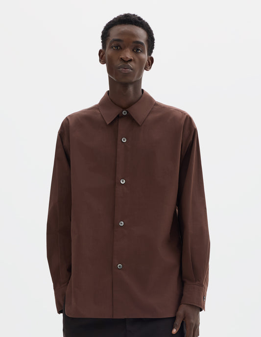 Cropped body image, front view of model wearing relaxed brown cotton shirt with mother of pearl buttons on wide stitch-through placket and curved hem. Styled with dark brown trousers.