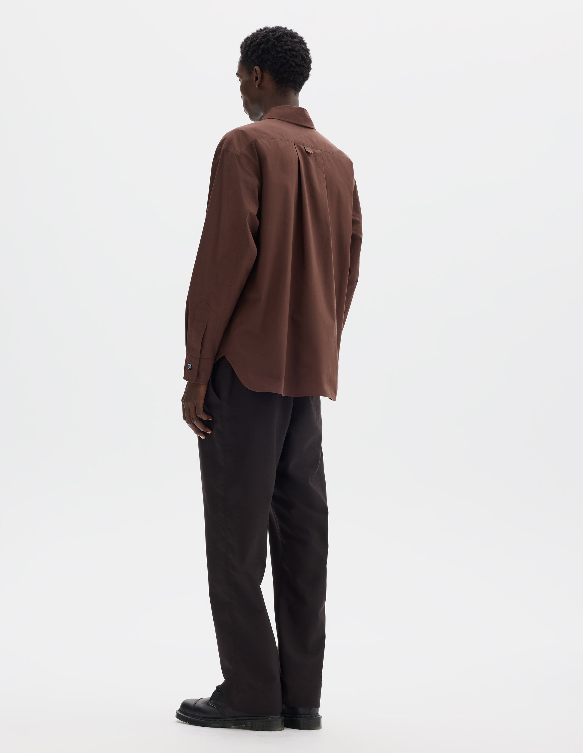 Full body image, back view of model wearing relaxed brown cotton shirt with split yoke and hanger loop detail on back, mother of pearl buttons on cuffs and curved hem. Styled with dark brown trousers and black leather shoes.
