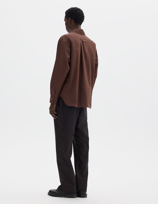 Full body image, back view of model wearing relaxed brown cotton shirt with split yoke and hanger loop detail on back, mother of pearl buttons on cuffs and curved hem. Styled with dark brown trousers and black leather shoes.