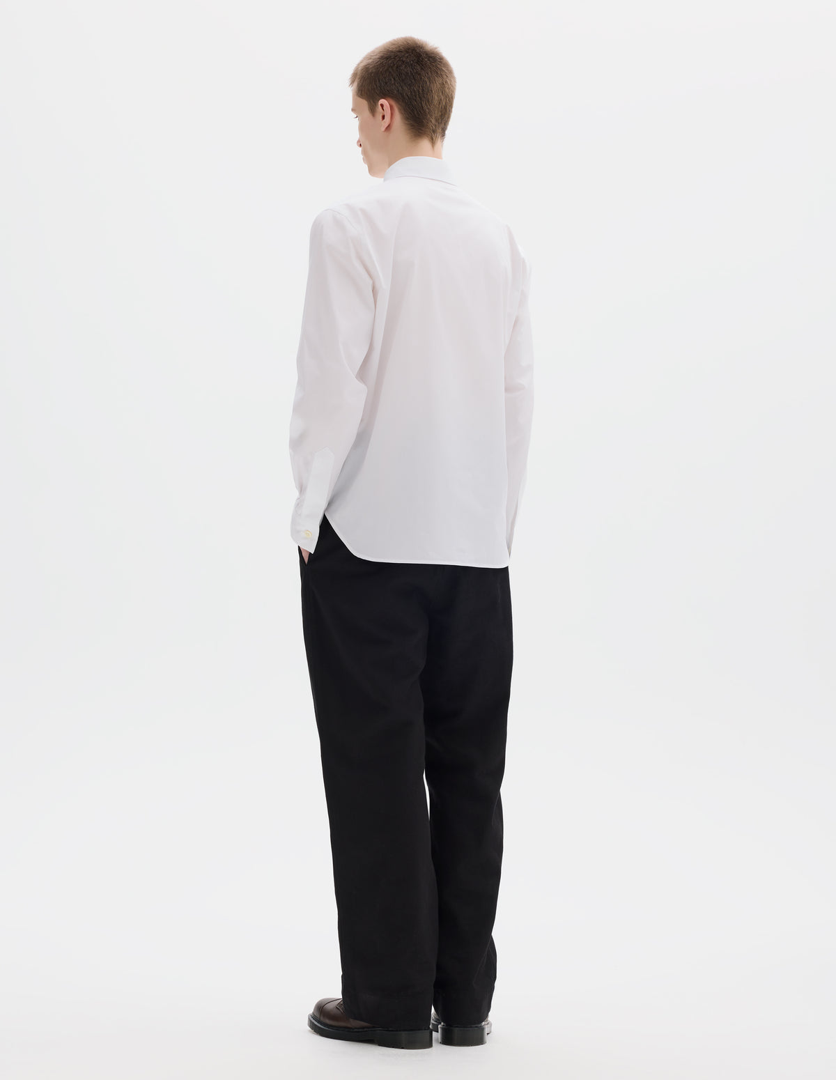 Full body image, back view of model wearing white cotton shirt with mother of pearl buttons on cuffs and curved hem. Styled with black trousers and brown leather shoes.