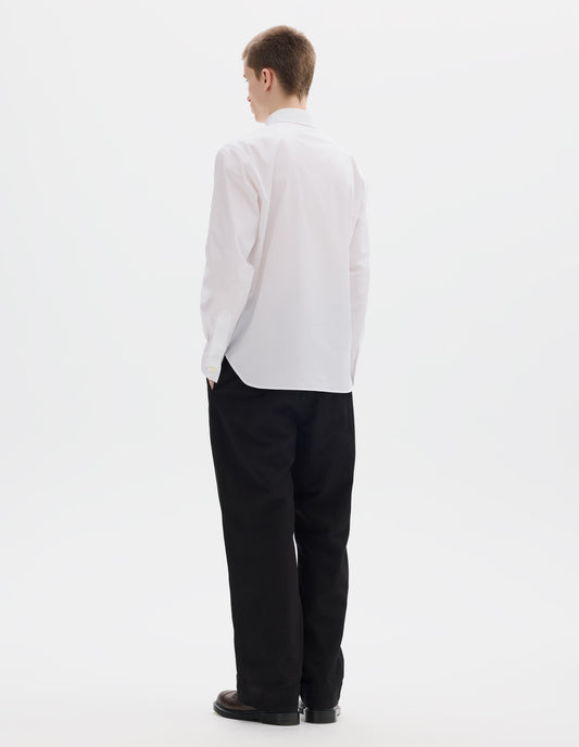 Full body image, back view of model wearing white cotton shirt with mother of pearl buttons on cuffs and curved hem. Styled with black trousers and brown leather shoes.