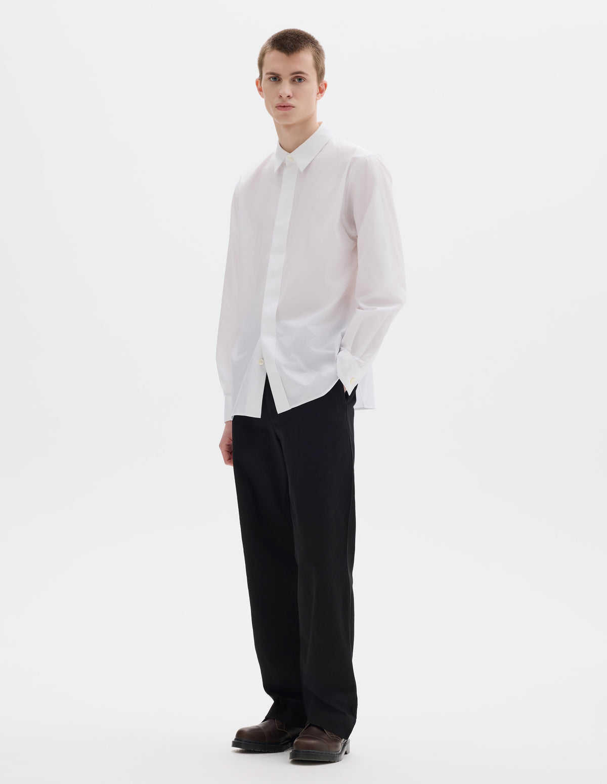 Full body image, front view of model wearing white cotton shirt with enclosed placket and mother of pearl button on collar, hanger loop on back neck and curved hem. Styled with black trousers and brown leather shoes.