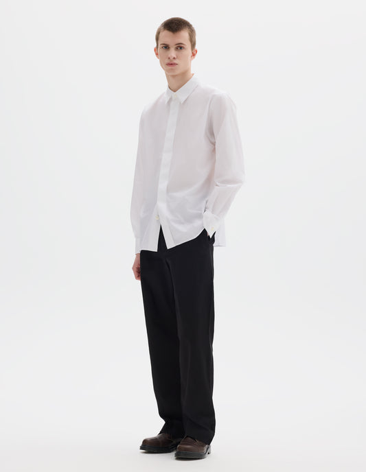 Full body image, front view of model wearing white cotton shirt with enclosed placket and mother of pearl button on collar, hanger loop on back neck and curved hem. Styled with black trousers and brown leather shoes.