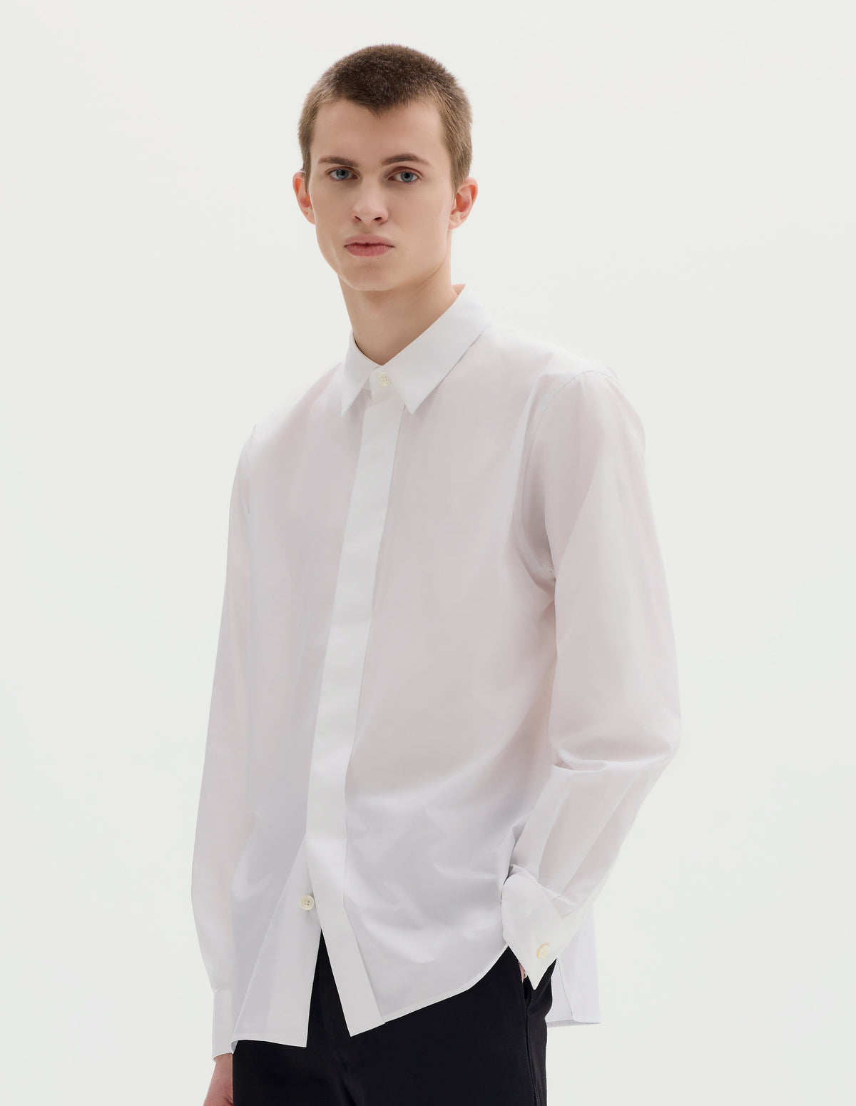Cropped body image, front view of model wearing white cotton shirt with enclosed placket and mother of pearl button on collar, hanger loop on back neck and curved hem. Styled with black trousers.
