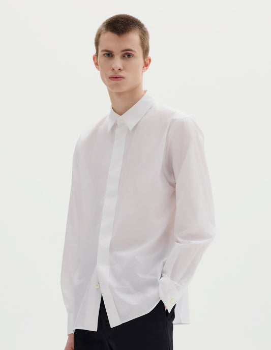 Cropped body image, front view of model wearing white cotton shirt with enclosed placket and mother of pearl button on collar, hanger loop on back neck and curved hem. Styled with black trousers.
