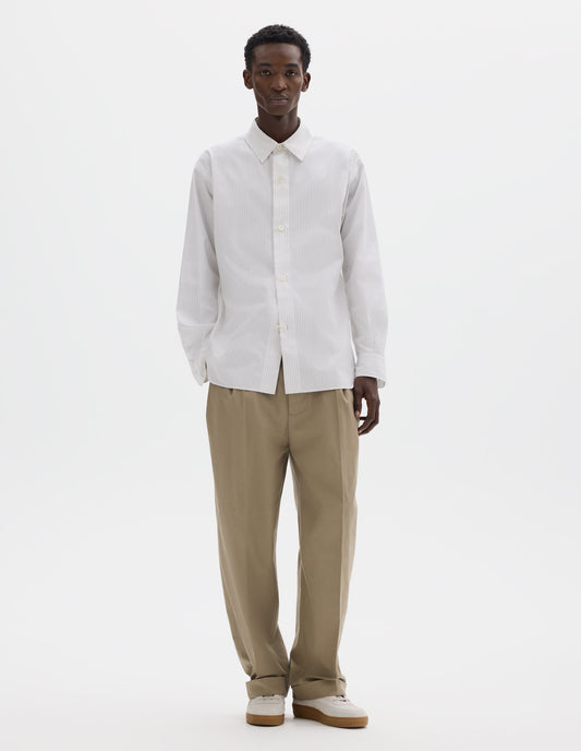 Full body image, front view of model wearing  relaxed white cotton shirt with green stripes, mother of pearl buttons on wide stitch-through placket and curved hem. Styled with dark beige trousers and beige nubuck trainers