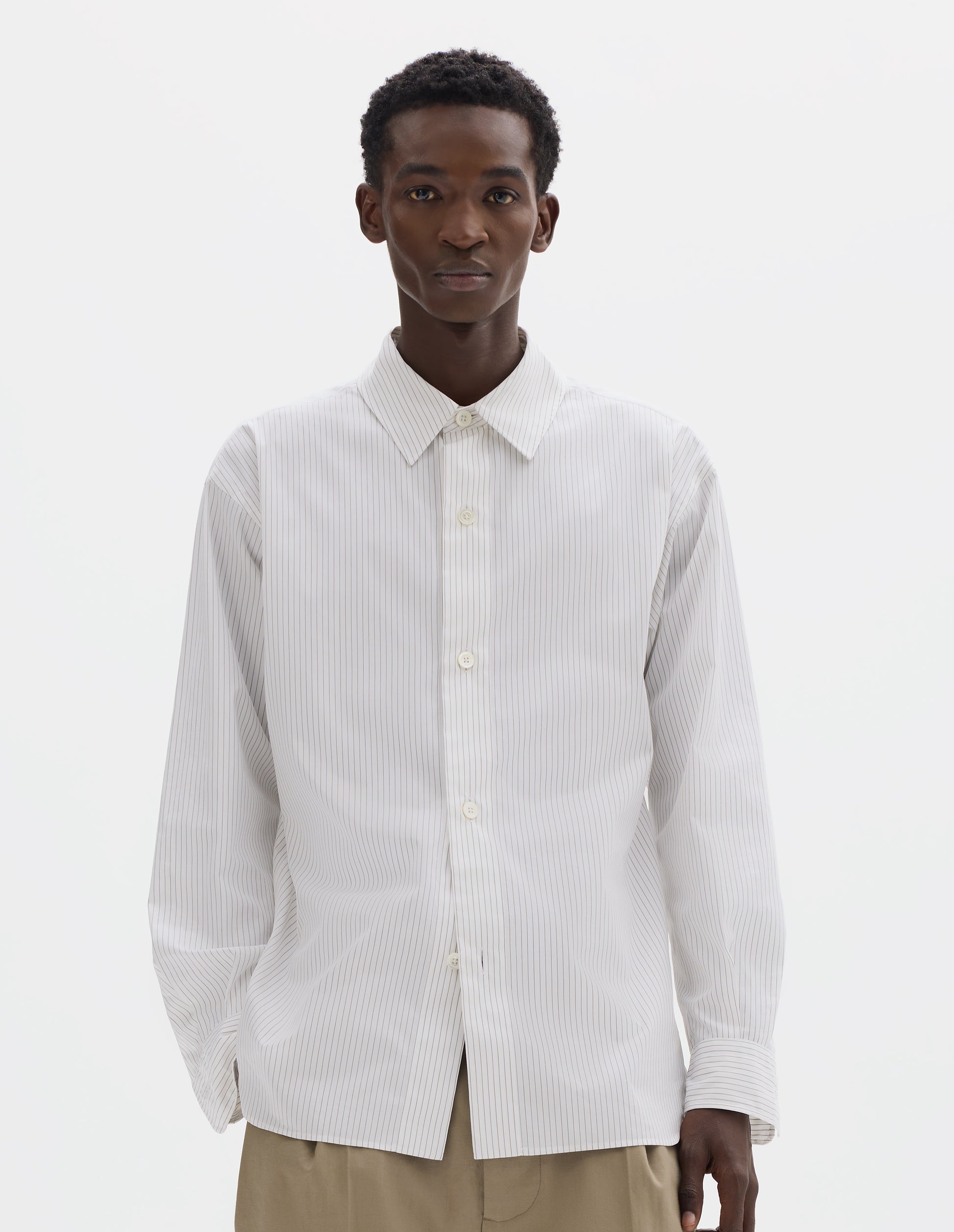 Cropped body image, front view of model wearing relaxed white cotton shirt with green stripes, mother of pearl buttons on wide stitch-through placket and curved hem. Styled with dark beige trousers.