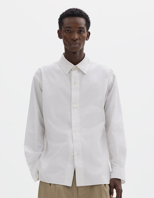 Cropped body image, front view of model wearing relaxed white cotton shirt with green stripes, mother of pearl buttons on wide stitch-through placket and curved hem. Styled with dark beige trousers.