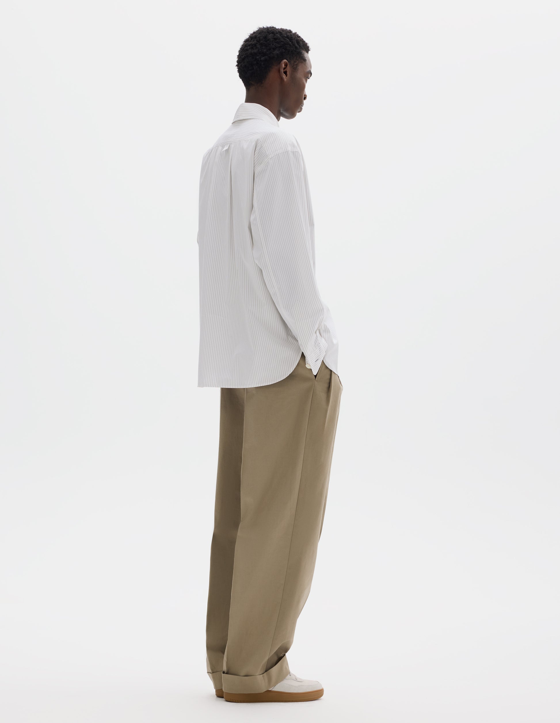 Full body image, back view of model wearing relaxed white cotton shirt with green stripes, split yoke with hanger loop detail on back, contrast cuffs with mother of pearl buttons in self fabric and curved hem. Styled with dark beige trousers and beige nubuck trainers