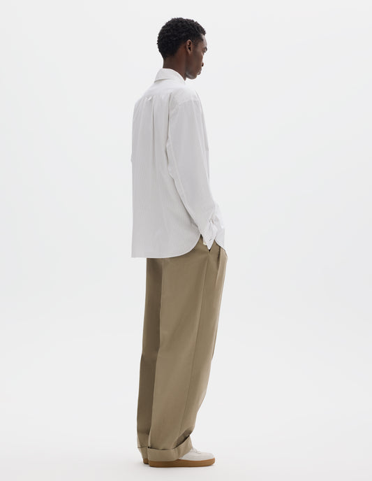 Full body image, back view of model wearing relaxed white cotton shirt with green stripes, split yoke with hanger loop detail on back, contrast cuffs with mother of pearl buttons in self fabric and curved hem. Styled with dark beige trousers and beige nubuck trainers