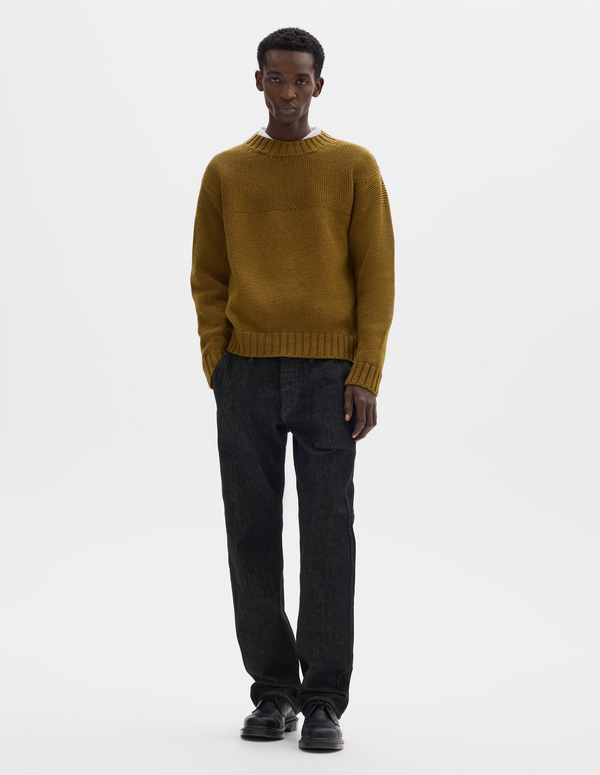 Full body image, front view of model wearing dark yellow relaxed wool jumper with set in sleeves, contrast garter stitch on yoke, and wide rib detailing on neck, cuffs and hem. Styled over white shirt, and with black jeans and black leather shoes.
