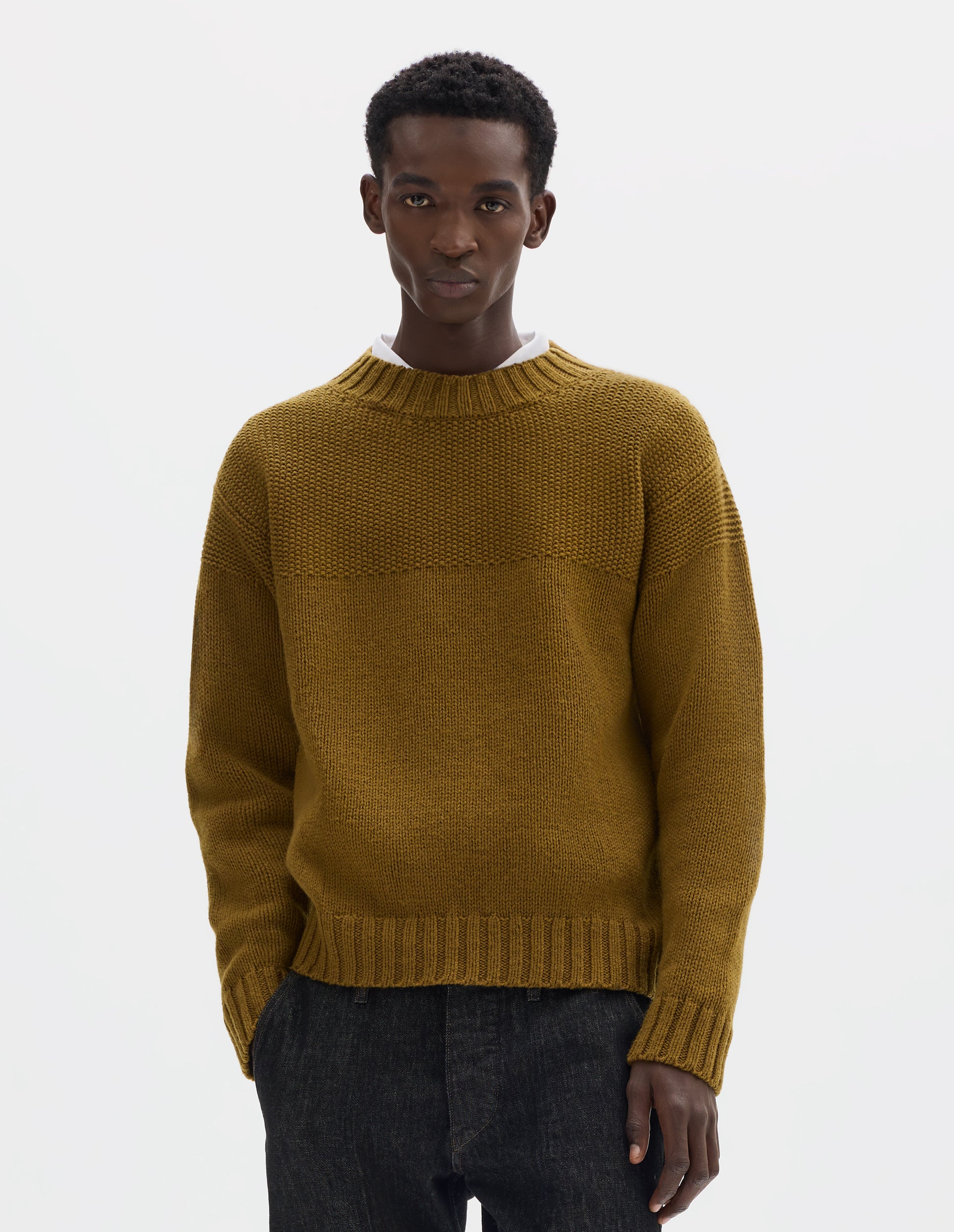 Cropped body image, front view of model wearing dark yellow relaxed wool jumper with set in sleeves, contrast garter stitch on yoke, and wide rib detailing on neck, cuffs and hem. Styled over white shirt and with black jeans.