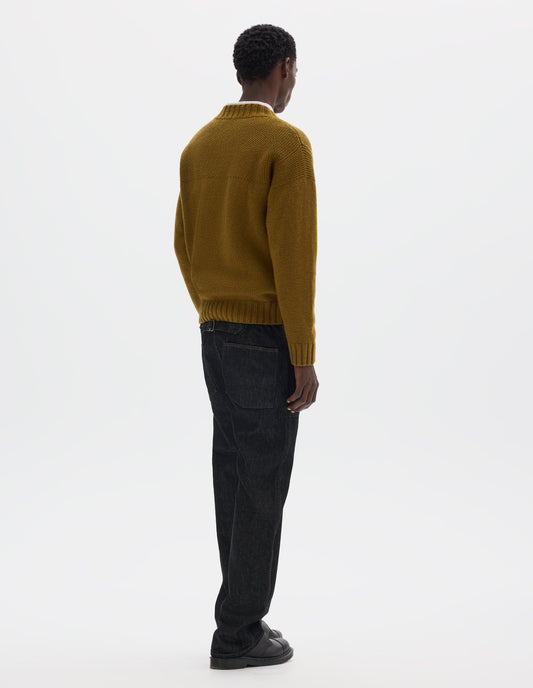 Full body image, back view of model wearing dark yellow relaxed wool jumper with set in sleeves, contrast garter stitch on yoke, and wide rib detailing on neck, cuffs and hem. Styled over white shirt, and with black jeans and black leather shoes.