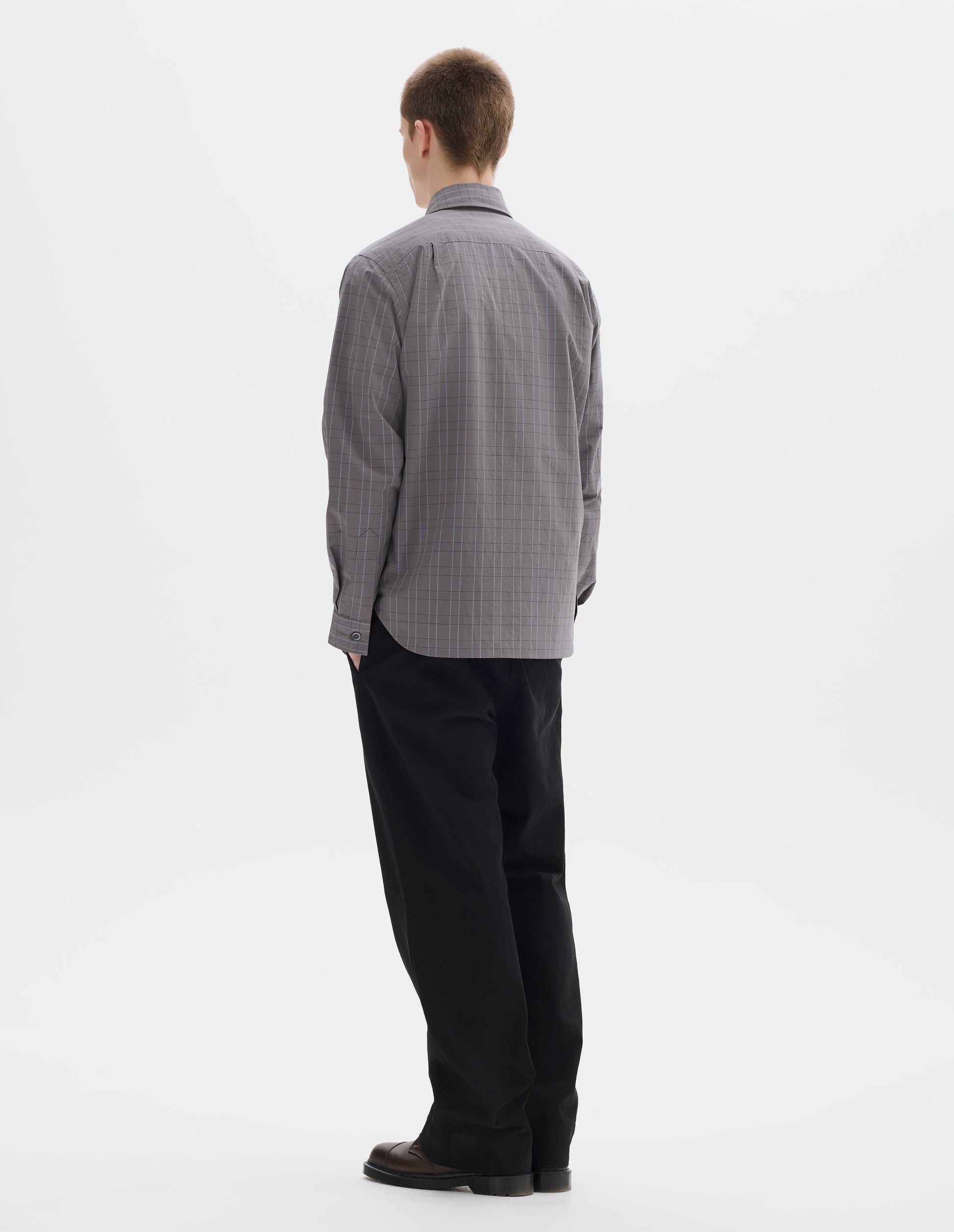 Full body image, back view of model wearing grey cotton shirt with dark grey and light blue checks, mother of pearl buttons on cuffs, curved hem and pleats on back yoke. Styled with black trousers and brown leather shoes.