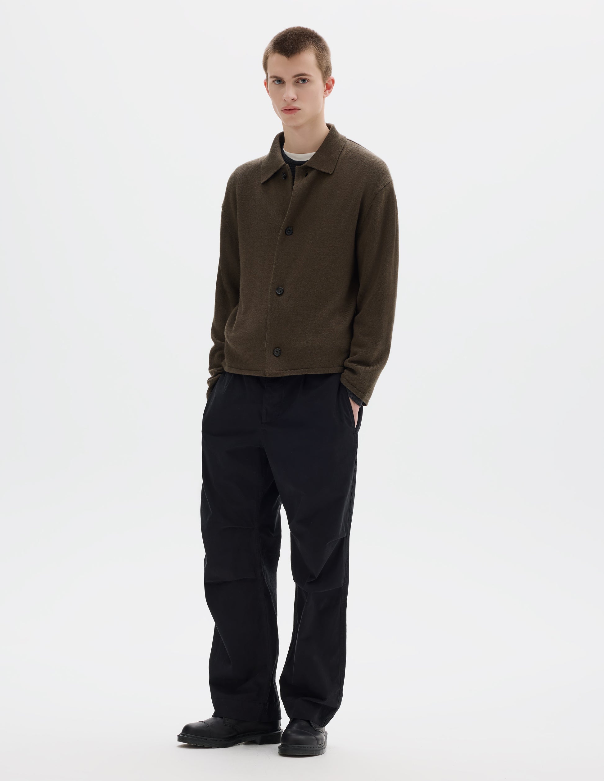 Full body image, front view of model wearing relaxed green merino cardigan with jersey stitch, fully fashioned collar, horn button fastening, set-in fully fashioned sleeves, and tubular hem and cuffs. Styled over white t-shirt and grey jumper, and with black trousers and black leather shoes.