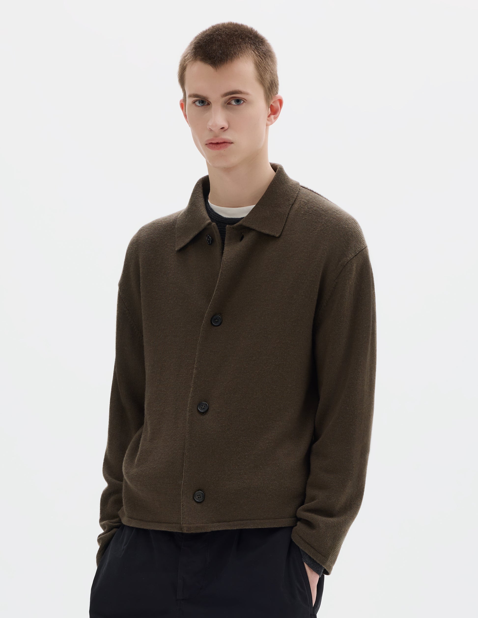 Cropped body image, front view of model wearing relaxed green merino cardigan with jersey stitch, fully fashioned collar, horn button fastening, set-in fully fashioned sleeves, and tubular hem and cuffs. Styled over white t-shirt and grey jumper, and with black trousers.