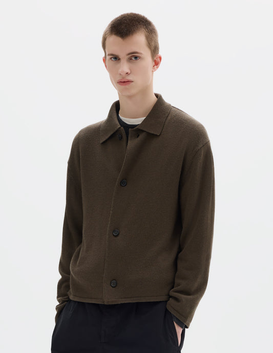 Cropped body image, front view of model wearing relaxed green merino cardigan with jersey stitch, fully fashioned collar, horn button fastening, set-in fully fashioned sleeves, and tubular hem and cuffs. Styled over white t-shirt and grey jumper, and with black trousers.