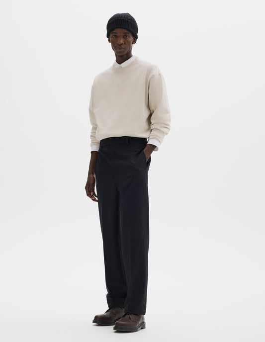 Full body image, front view of model wearing relaxed dark blue wool cotton trousers with tapered leg, belt loops and waistband extension with hook and bar fastening. Styled with black beanie, white shirt, beige jumper and brown leather shoes.
