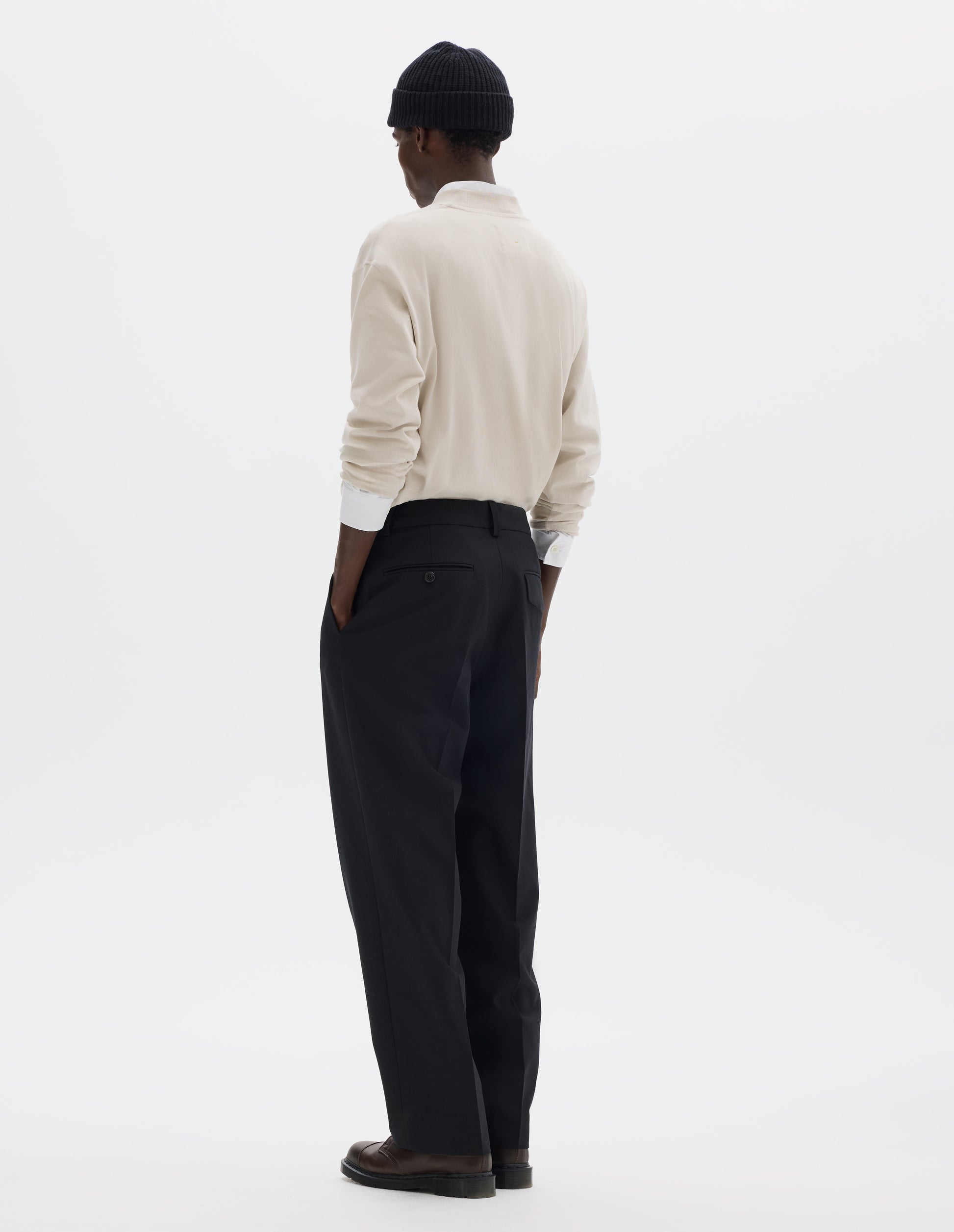 Full body image, back view of model wearing relaxed dark blue wool cotton trousers with tapered leg, belt loops, and with one jetted flap back pocket and one horn button through jetted back pocket. Styled with dark blue beanie, white shirt, white long sleeve t-shirt and brown leather shoes.