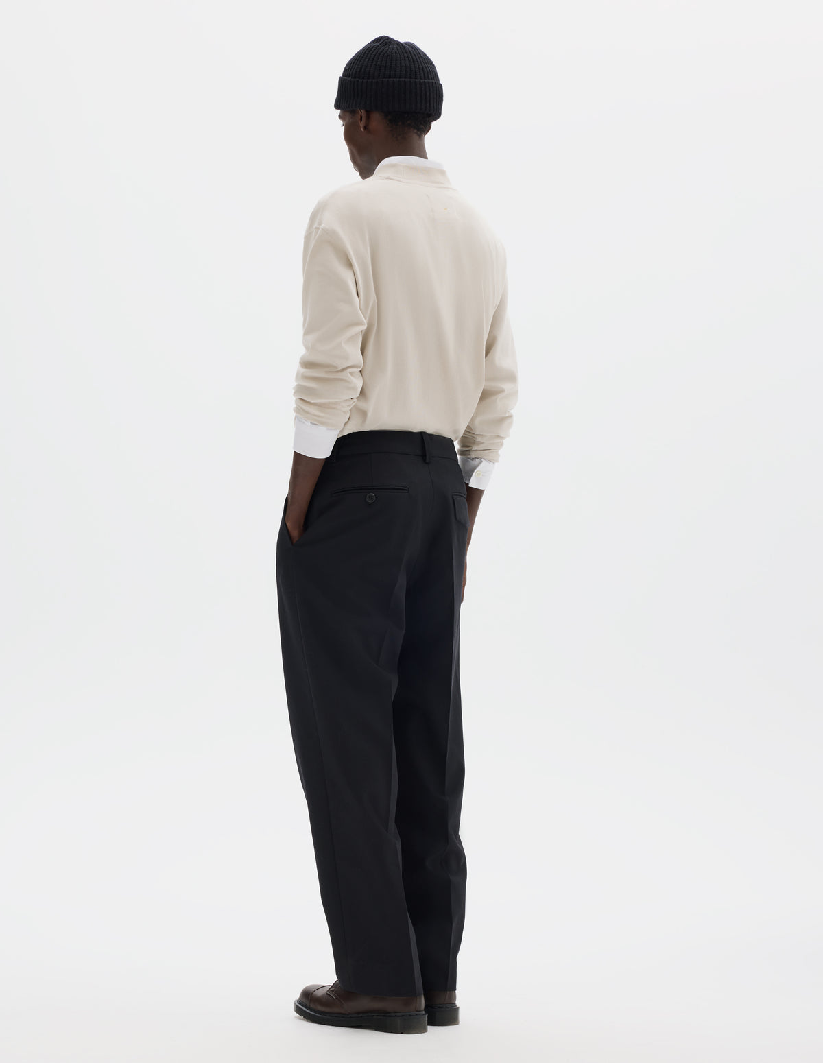 Full body image, back view of model wearing relaxed dark blue wool cotton trousers with tapered leg, belt loops, and with one jetted flap back pocket and one horn button through jetted back pocket. Styled with black beanie, white shirt, beige jumper and brown leather shoes.