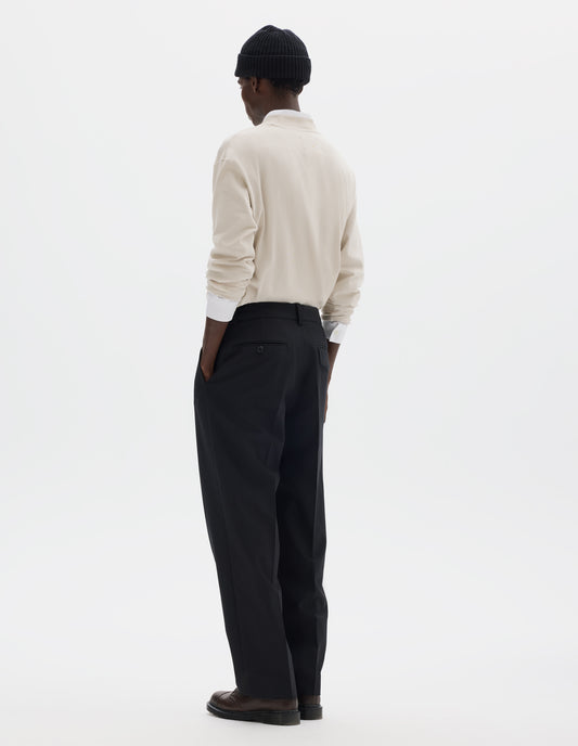 Full body image, back view of model wearing relaxed dark blue wool cotton trousers with tapered leg, belt loops, and with one jetted flap back pocket and one horn button through jetted back pocket. Styled with black beanie, white shirt, beige jumper and brown leather shoes.