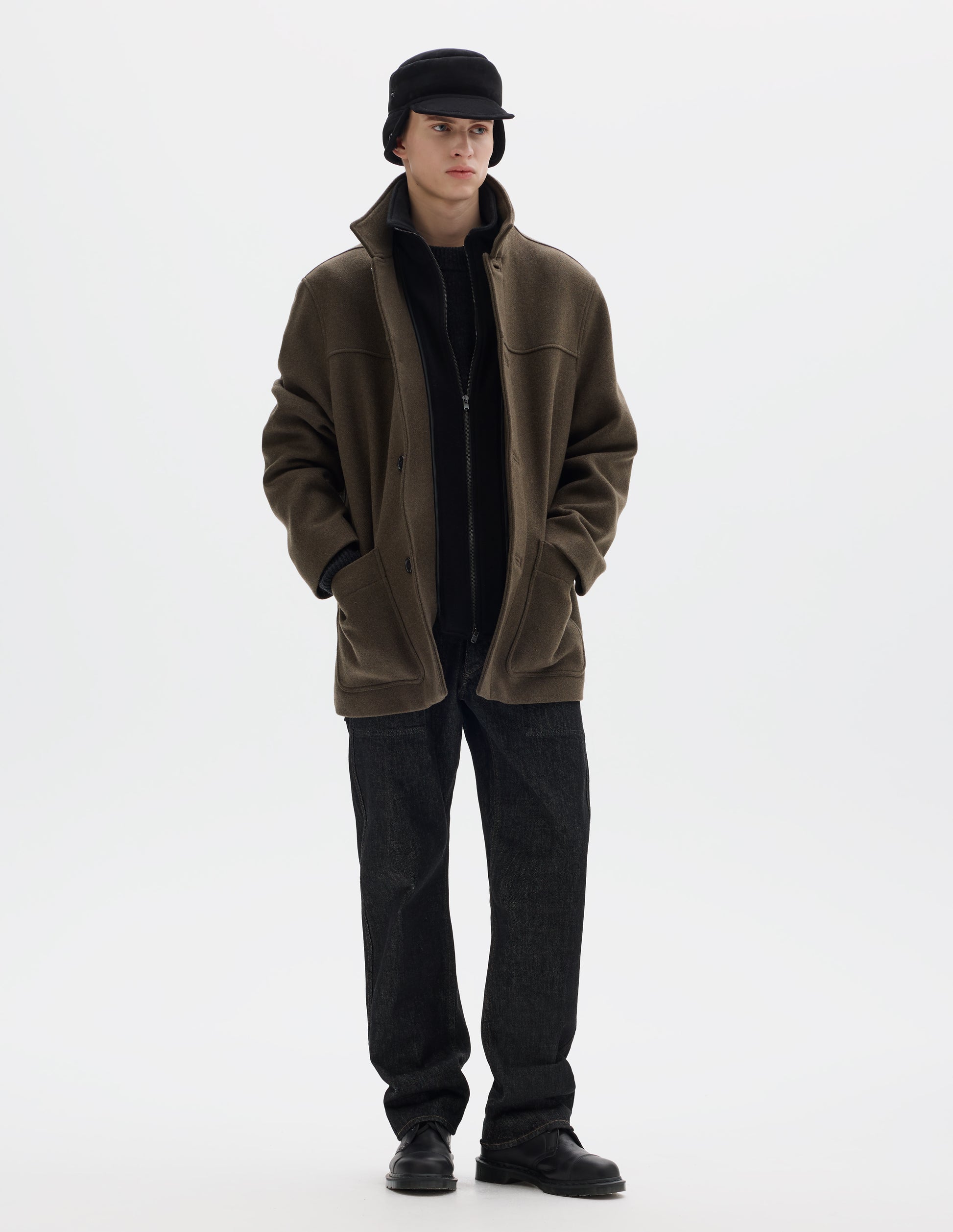 Full body image, front view of model wearing khaki green relaxed wool coat with internal funnel collar with zip closure and detachable fleece facing, external half mock collar, front yoke, horn buttons and jetted buttonholes, and patch pockets. Styled open with black shearling cap, black jeans and black leather shoes. 