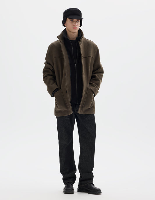 Full body image, front view of model wearing khaki green relaxed wool coat with internal funnel collar with zip closure and detachable fleece facing, external half mock collar, front yoke, horn buttons and jetted buttonholes, and patch pockets. Styled open with black shearling cap, black jeans and black leather shoes. 