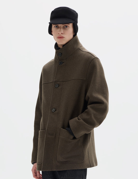 Cropped body image, front view of model wearing khaki green relaxed wool coat with half mock collar, horn buttons and jetted buttonholes, front yoke and patch pockets. Styled buttons done up and with black shearling cap and black jeans.