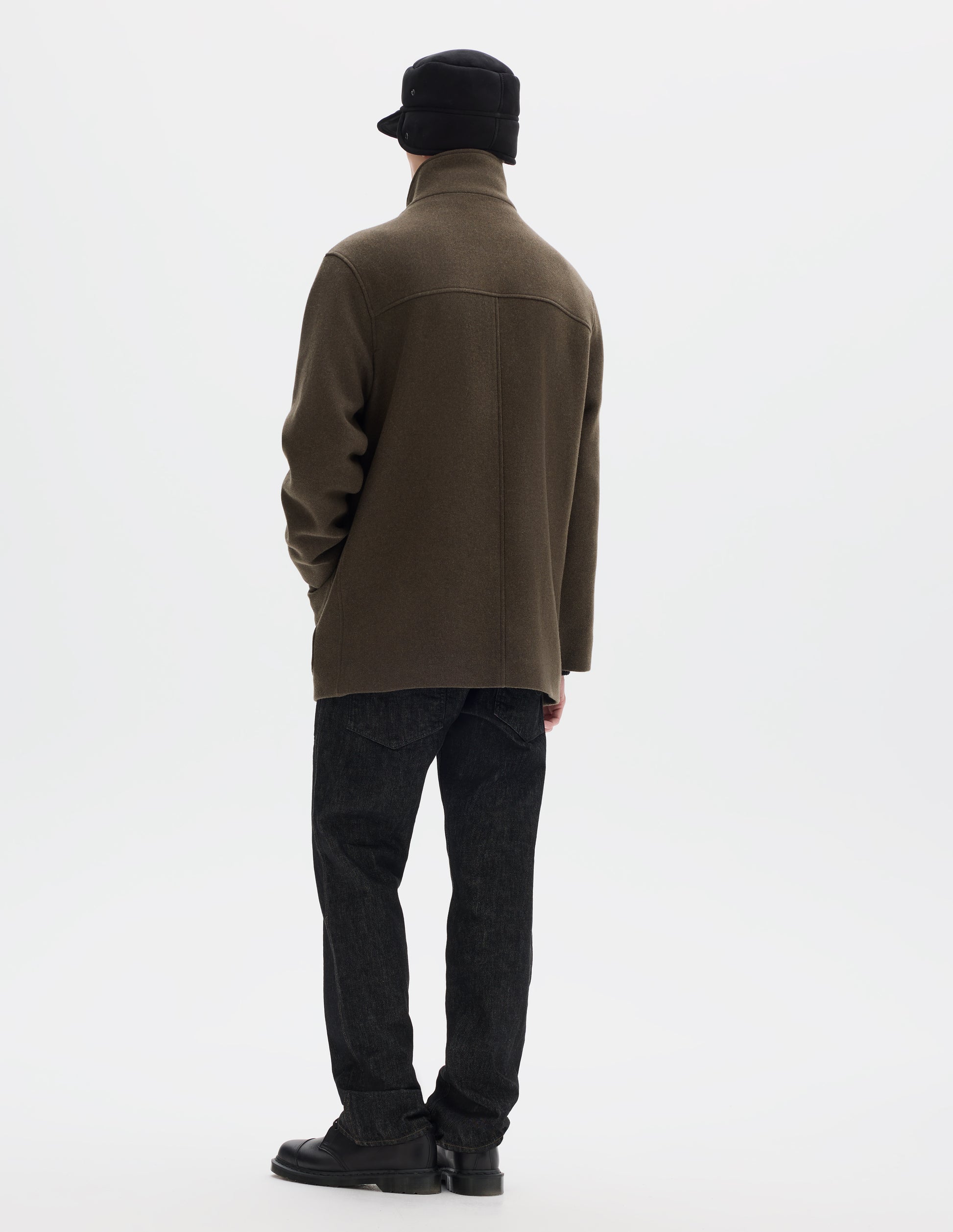 Full body image, back view of model wearing khaki green relaxed wool coat with half mock collar, back yoke and centre back seam. Styled with black shearling cap, black jeans and black leather shoes. 