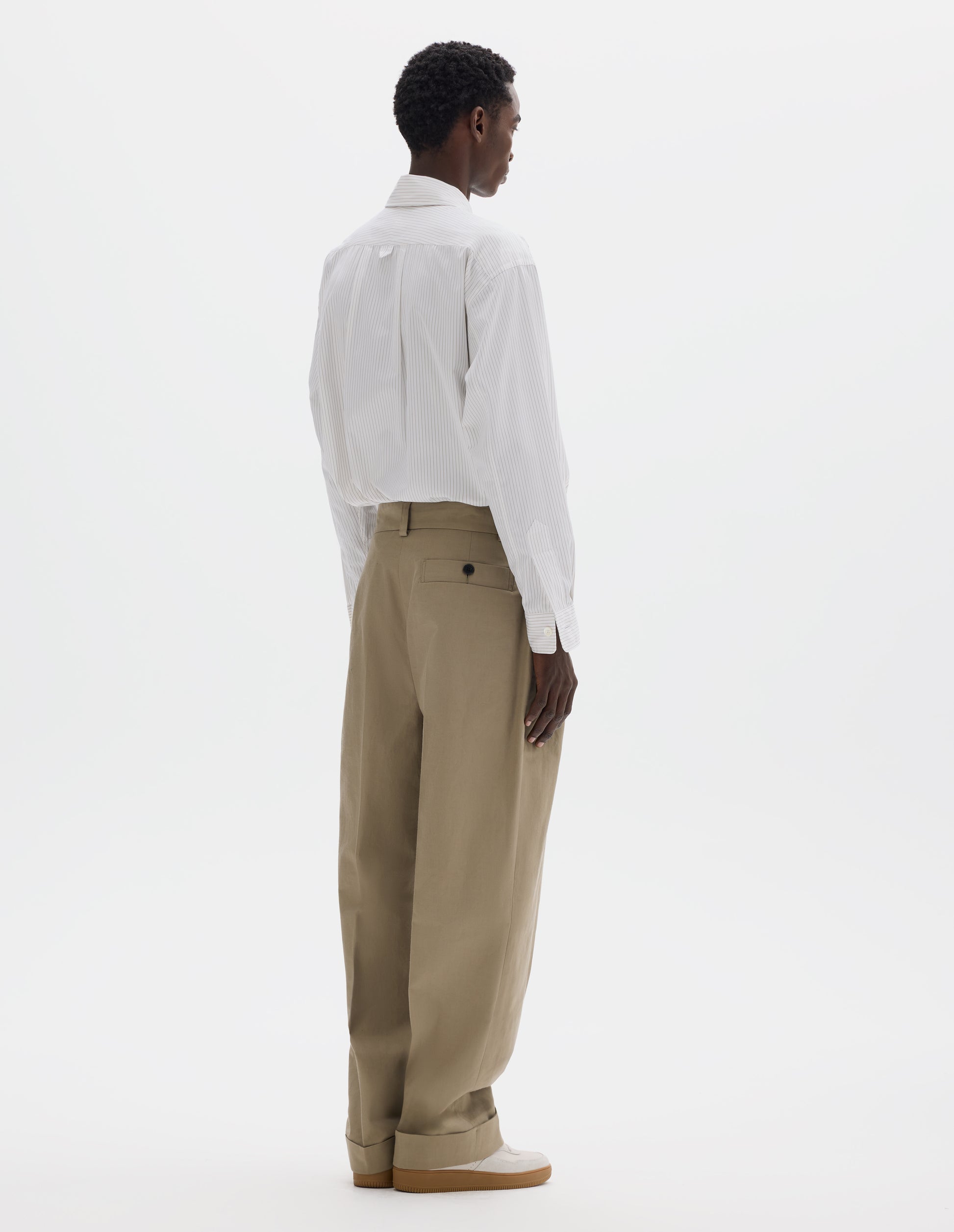 Full body image, back view of model wearing relaxed beige cotton pleated trousers with straight leg, belt loops and deep horn button-through welt back pocket. Styled with striped white shirt and light beige nubuck trainers.