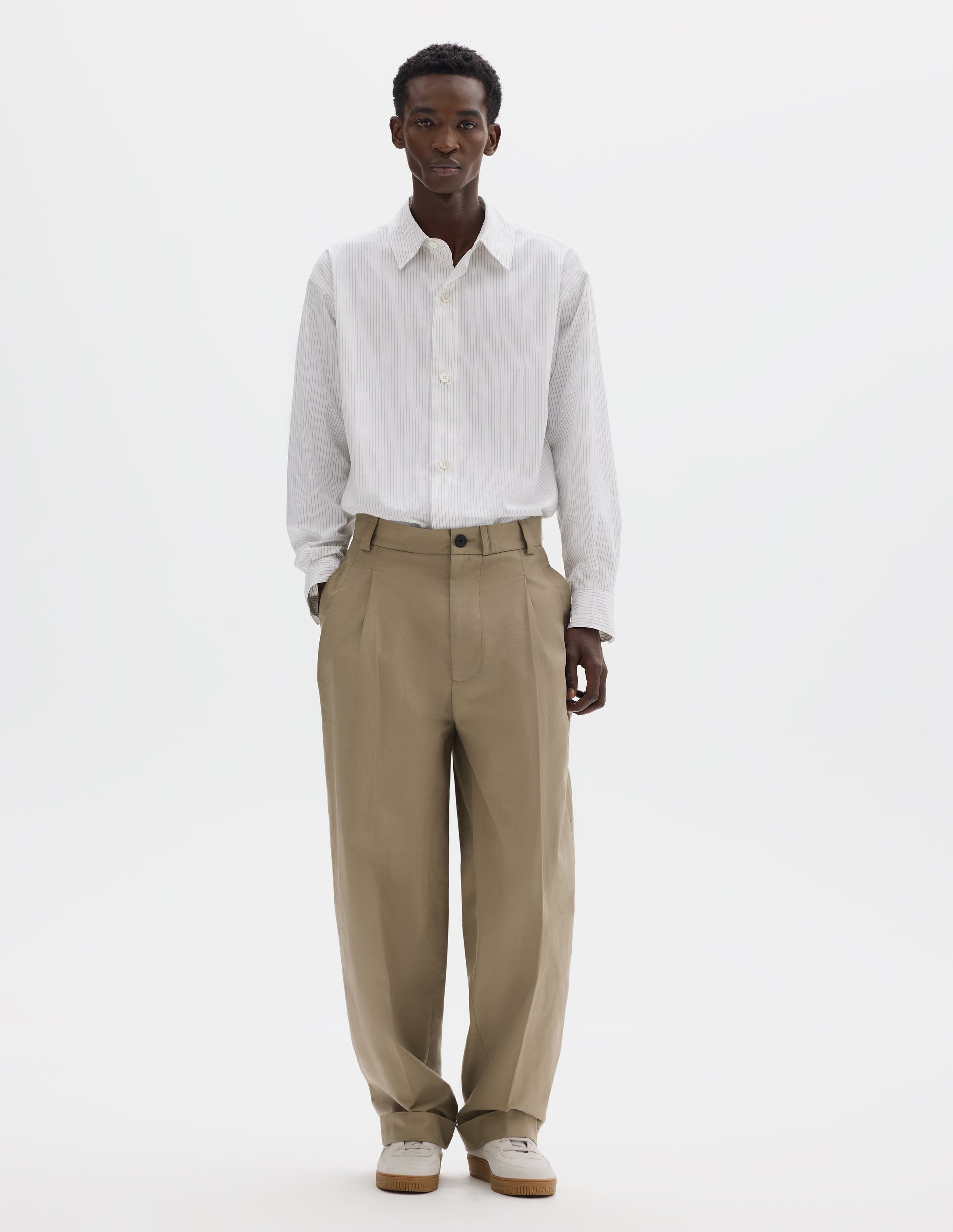 Full body image, front view of model wearing relaxed beige cotton pleated trousers with straight leg, horn button-through waistband with belt loops, concealed side pockets and cuffed hem. Styled with striped white shirt and light beige nubuck trainers.