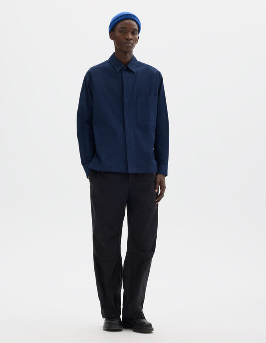 Full body image, front view of model wearing dark blue relaxed cotton shirt with concealed placket and mother of pearl button on collar, large patch chest pocket with internal pen pocket and vent split hem. Styled with blue hat, black trousers and black leather shoes.