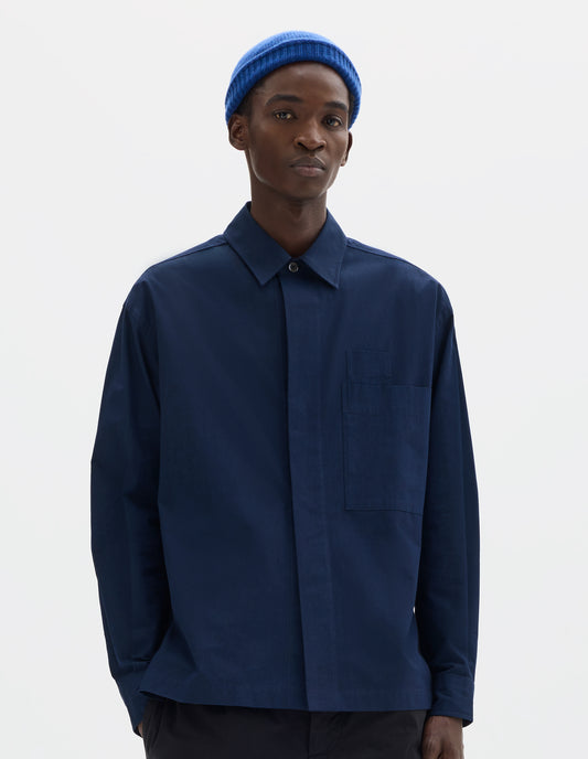 Cropped body image, front view of model wearing dark blue relaxed cotton shirt with concealed placket and mother of pearl button on collar, large patch chest pocket with internal pen pocket and vent split hem. Styled with blue hat and black trousers.