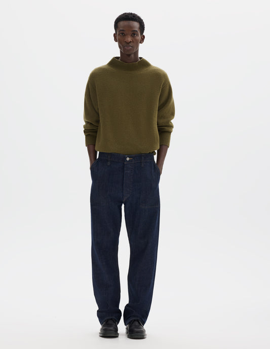 Full body image, front view of model wearing indigo denim twill jeans, with metal button fly, grown on waistband with belt loops and stitch-through slant pockets. Styled with olive green mock neck sweater and black leather shoes.