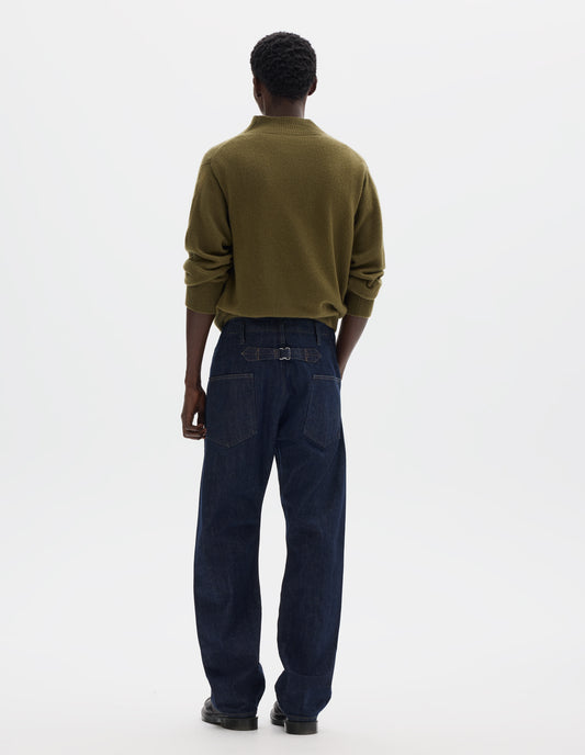 Full body image, back view of model wearing indigo denim twill jeans with grown on waistband with belt loops and back pockets with half cotton lining, cinch on back. Styled with olive green mock neck sweater and black leather shoes.