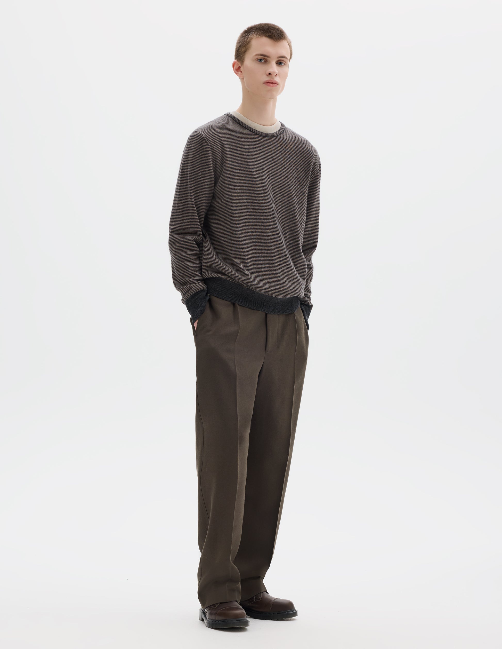 Full body image, front view of model wearing grey merino cashmere jumper with dark grey stripes, crew neck with tubular collar, and with contrast rib on cuffs and hem. Styled over white t-shirt and with khaki green trousers and brown leather shoes.