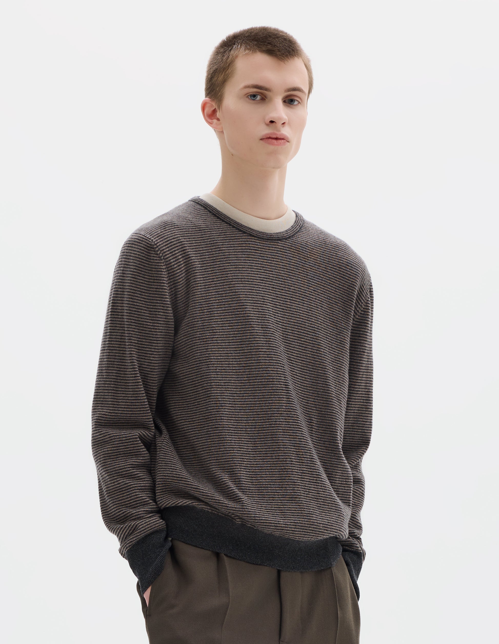Cropped body image, front view of model wearing grey merino cashmere jumper with dark grey stripes, crew neck with tubular collar, and with contrast rib on cuffs and hem. Styled over white t-shirt and with khaki green trousers.