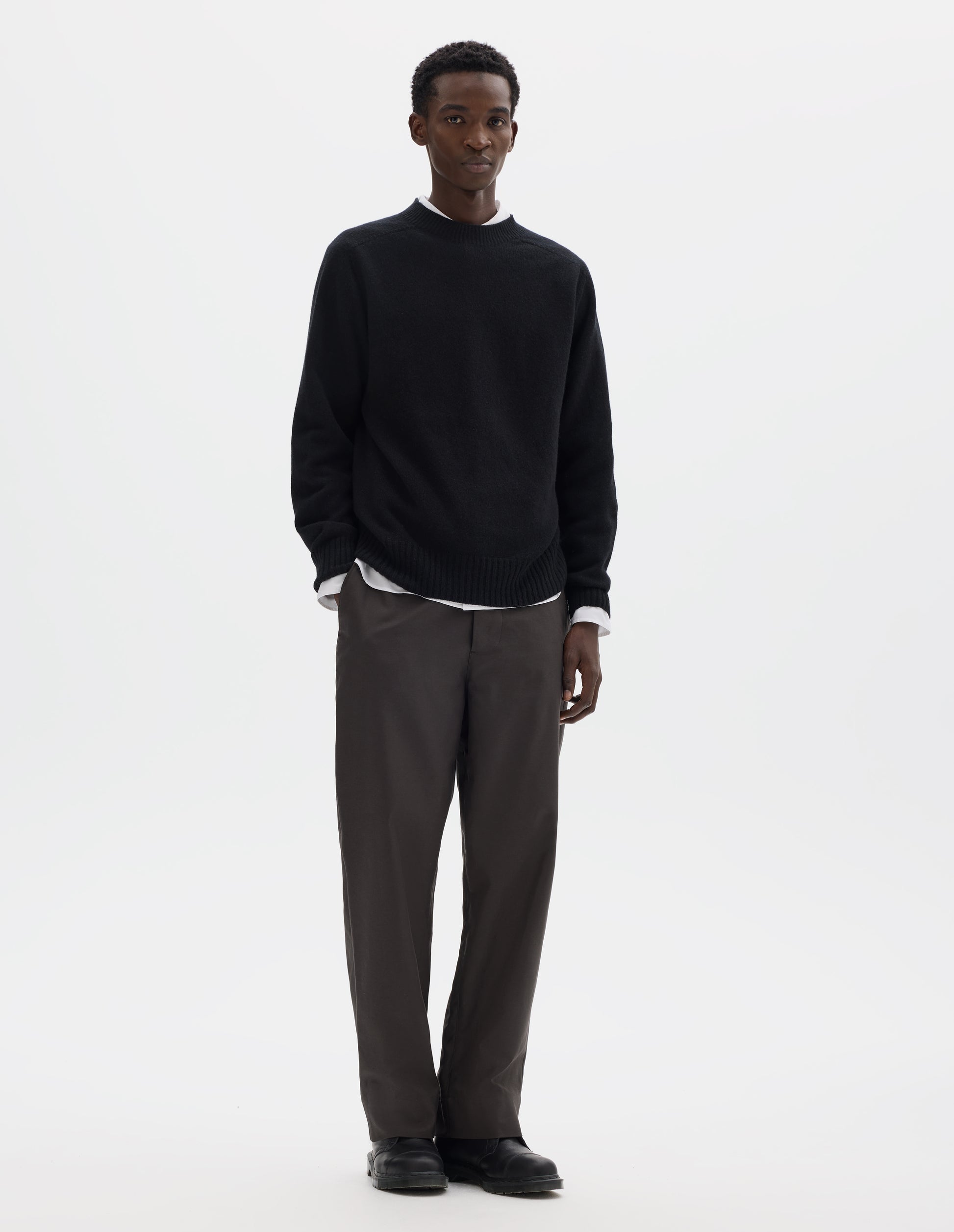 Full body image, front view of model wearing black merino cashmere seamless jumper with ribbed crew neck, cuffs and hem. Styled over white shirt, and with grey trousers and black leather shoes.